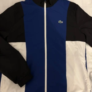 Blå och svart vindjacka från Lacoste - Säljer en stilren blå och svart vindjacka från Lacoste med dragkedja framtill. Jackan har långa ärmar och det ikoniska Lacoste-logotypen på bröstet och ryggen. Perfekt för en sportig look!