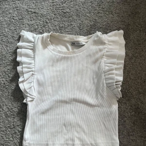 Vit topp från Zara - Storlek S. Säljer en söt vit ribbad topp från Zara med volangdetaljer på axlarna. Använd få gånger, fint skick.Pris går att diskutera!