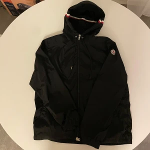 Svart vindjacka från Moncler - Säljer en stilren svart vindjacka från Moncler med dragkedja och huva. Jackan har Monclers logotyp på bröstet och en randig detalj på huvan. Perfekt för blåsiga dagar.