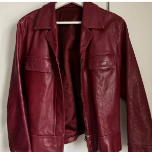 Vinröd skinnjacka - Snygg vinröd skinnjacka med klassisk krage och två bröstfickor. Jackan har en stilren design med långa ärmar och en rak passform. Perfekt för att ge din outfit en cool touch.