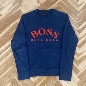Mörkblå tröja från Hugo Boss - Säljer en stilren mörkblå tröja från Hugo Boss med röd/orange logotyp på framsidan. Tröjan är perfekt för en avslappnad men ändå snygg look. 
