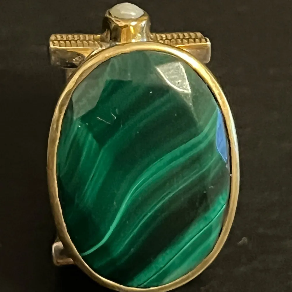 Unik ring från Canyon med en stor oval grön sten i centrum, omgiven av guldfärgad infattning. Sidorna pryds av små pärldetaljer och ett intrikat mönster på bandet. Perfekt för att ge en elegant touch till din stil.. Asusteet.