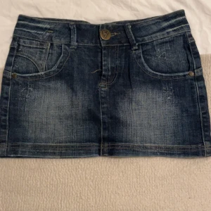 Jeanskjol med fickor - Snygg jeanskjol i mörkblå denim med slitna detaljer och dekorativa sömmar på bakfickorna. Kjolen har en klassisk femficksdesign och stängs med en knapp och dragkedja framtill. Perfekt för en avslappnad stil.