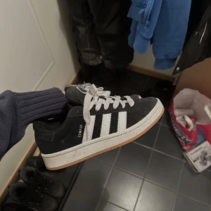 Svarta Adidas Campus sneakers - Säljer ett par svarta Adidas Campus sneakers med vita ränder och snörning. Skorna har en klassisk design med en vit sula och är perfekta för en avslappnad stil. Dom är fåtal använda och tvättas såklart innan frakten. 
