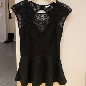 Svart klänning med spetsdetaljer från Gina Tricot - Snygg svart topp från Gina Tricot med spetsdetaljer på axlar och över bröstet. Toppen har en elegant design med en liten volang nedtill och även öppen vid bröstet. Perfekt för en kväll ute eller en speciell tillställning.