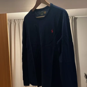 Mörkblå långärmad tröja från Ralph Lauren - Säljer en stilren mörkblå långärmad tröja från Ralph Lauren med det klassiska röda logotypen på bröstet. Perfekt för en avslappnad men ändå elegant look. Passar bra till både jeans och chinos.