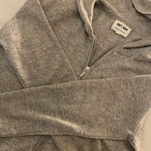 Grå hoodie från Soft Goat - Mysig grå hoodie från Soft Goat med dragkedja framtill. Tillverkad i mjukt material med långa ärmar och en praktisk huva. 100 % cashmere. Perfekt för kyliga dagar.