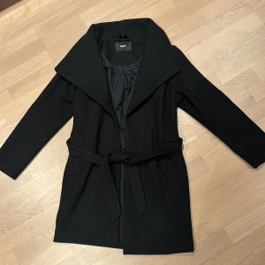 Svart kappa från Bik Bok - Elegant svart kappa från Bik Bok i storlek XS. Tillverkad av en akryl- och polyesterblandning med ett bälte i midjan för en snygg siluett. Perfekt för kyligare dagar.