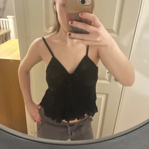 Svart linne med spetsdetaljer - Säljer ett svart linne med smala axelband och spetsdetaljer. Toppen har en v-ringad design och en volangkant nedtill. Perfekt för en stilren och elegant look.