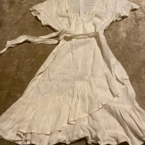 Vit omlottklänning med volang - Charmig vit omlottklänning med volangdetaljer och knytband i midjan. Klänningen har korta ärmar och en v-ringad halslinje!🤍