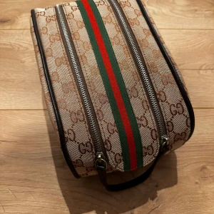 Beige necessär  - Snygg beige necessär  med det klassiska GG-mönstret. Den har en grön och röd rand i mitten och två dragkedjor upptill. Perfekt för att hålla ordning på dina saker med stil.