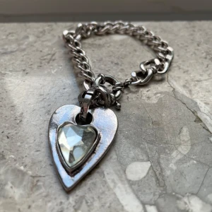 Silverfärgat armband med hjärthänge - Stilrent silverfärgat armband med ett hjärthänge. Hänget har en glittrig sten på framsidan och ingraverad text på baksidan. Perfekt för att ge en elegant touch till din outfit. Obs finns små defekter som rost, dock går det att få bort enkelt och syns knappt. Aldrig använd!