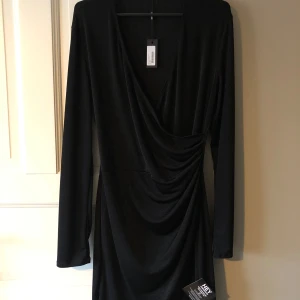 Svart omlottklänning från ICHI - Elegant svart omlottklänning från ICHI med långa ärmar och draperad design. Perfekt för en stilren look. Lite glansig/glittrig i materialet, aldrig kommit till användning