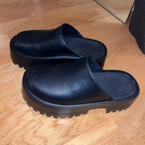 Svarta platåskor - Säljer ett par svarta platåskor med grov sula. Skorna har en stilren design utan snörning och är tillverkade i syntetmaterial. Perfekta för att ge lite extra höjd och stil till din outfit.