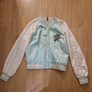 Ljusblå och vit bomberjacka med broderi - Snygg ljusblå och vit bomberjacka med broderade detaljer på bröstet och ryggen. Jackan har långa ärmar och stängs med knappar framtill. Perfekt för en cool och avslappnad stil.