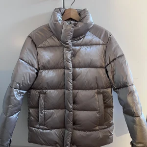 Silverfärgad pufferjacka från MNG - Snygg och glansig silverfärgad pufferjacka från MNG. Jackan har en hög krage och dragkedja framtill. Perfekt för kyliga dagar med sin vattenavvisande yta.