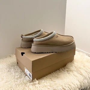 UGG tazz tasman tofflor - Säljer ett par ljusbeiga UGG Tasman med plattform och ljusblå detalj som använts endast vid 1-2 tillfällen. Därav är de i nyskick, vilket även syns på sulan. Tycker de är otroligt snygga men kommer tyvärr inte till användning. Väldigt bekväma och praktiska för att göra snabba ärenden samt snygga att ha i vardagen. Funkar både inomhus och utomhus. Storlek 39 men passar perfekt för mig som har storlek 38. Dessa kommer med kartong.