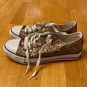 Glittriga sneakers med snörning - Snygga sneakers i guld med glittrig yta och vita snören. Skorna har en klassisk design med vit tå och sula, perfekt för att ge en extra touch till din outfit.