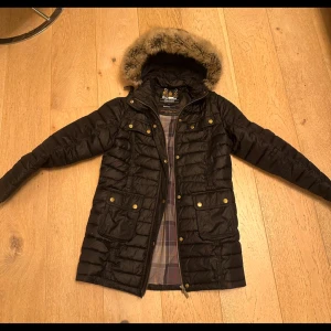 Svart dunjacka från Barbour - Snygg svart dunjacka från Barbour med pälskantad huva och dragkedja. Jackan har flera praktiska fickor med knappar och ett klassiskt rutigt foder. Perfekt för kyliga dagar.