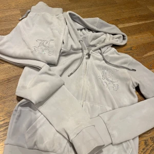 Grått set från Juicy Couture XS *som nytt* - Säljer en grå hoodie och matchande byxor från Juicy Couture. Setet är dekorerat med glittriga stenar som bildar texten 'Juicy' på både ryggen av hoodien och baksidan av byxorna. Perfekt för en avslappnad och stilren look. Tröjan använd en gång, byxan 3. Som nytt!!