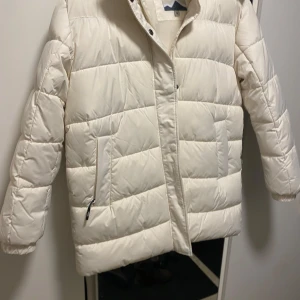 Vit pufferjacka från Tuxer - Säljer en snygg vit pufferjacka från Tuxer i storlek M. Jackan har en quiltad design med dragkedja och huva, perfekt för kyliga dagar. Den är både stilren och praktisk med fickor framtill. Använd 5 gånger helt i nyskick 