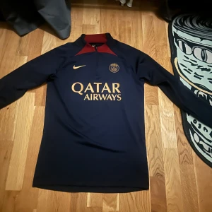 Mörkblå långärmad fotbollströja från Nike - Säljer en stilren mörkblå fotbollströja från Nike med PSG-logga och Qatar Airways-tryck i guld. Tröjan har en röd krage och är tillverkad i Dri-FIT-material för optimal komfort. Perfekt för fotbollsfans!