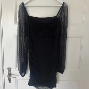 Svart klänning med transparenta ärmar - Elegant svart klänning med långa, transparenta ärmar och en tight passform. Klänningen har en stilren design med en fyrkantig halsringning och är perfekt för festliga tillfällen.