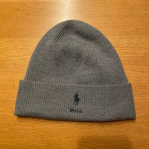 Grå ribbad mössa från Polo Ralph Lauren - Snygg grå ribbad mössa från Polo Ralph Lauren med broderad logga framtill. Perfekt för kyliga dagar och en stilren look. 10 av 10 skick Aldrig använd