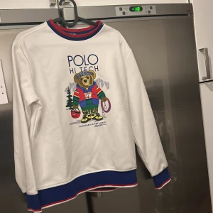 Vit tröja med Polo Bear från Ralph Lauren - Säljer en vit tröja från Ralph Lauren med det ikoniska Polo Bear-motivet. Tröjan har blå och röda detaljer vid krage och ärmslut. Perfekt för en avslappnad stil med en touch av lekfullhet. Stk L (14-16)