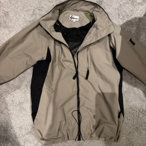 Beige och svart vindjacka - Snygg beige och svart vindjacka med dragkedja och huva. Perfekt för blåsiga dagar. Jackan har en praktisk ficka på ärmen och justerbar nederkant.