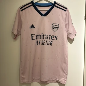 Rosa Arsenal fotbollströja från Adidas - Snygg rosa Arsenal fotbollströja från Adidas med korta ärmar och mörkblå detaljer. Tröjan har klubbens emblem på bröstet och sponsorlogga framtill. Perfekt för alla Arsenal-fans!