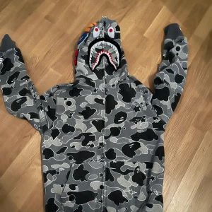 Grå camo hoodie från A Bathing Ape - Säljer en grå camo hoodie från A Bathing Ape med hajmotiv på huvan och färgglada bokstäver 'WGM' på ärmen. Hoodien har dragkedja och är långärmad, perfekt för en streetwear-look. Koftan är storlek L men passar perfekt för någon med storlek M som mig själv. Den sitter perfekt och man kan dra upp dragkedjan så koftan täcker ditt ansikte 