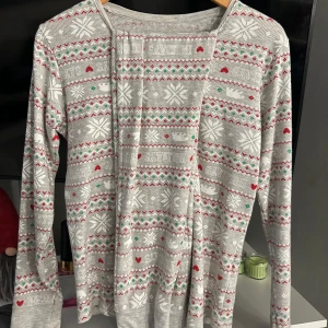 Grå julpyjamas - Säljer ett set med en grå långärmad tröja och matchande leggings. Båda plaggen har ett festligt julmönster med hjärtan och snöflingor i rött och grönt. Perfekt för mysiga vinterdagar! 🎄Har endast använt vid ett tillfälle