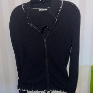 Vintage Guess Svart ribbad kofta  - Snygg svart kofta från Guess med dragkedja framtill några små noppror på framsidan.På etiketten hittar jag ingen storlek uppskattar till S-M, kan skicka foto på etiketten Den har en ribbad struktur och vita detaljer längs kanterna. Perfekt för en stilren look.