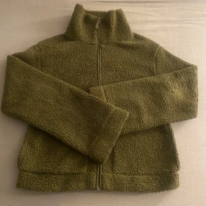 Olivgrön teddyjacka med dragkedja - Mysig olivgrön teddyjacka med dragkedja framtill. Perfekt för kyliga dagar med sin fluffiga och varma design. 