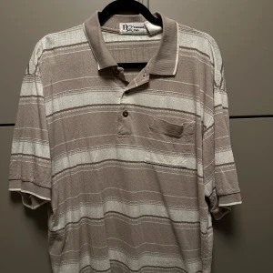 Beige och vit randig pikétröja från RQ Classics - Säljer en klassisk beige och vit randig pikétröja från RQ Classics. Tröjan har en bröstficka och knappar vid kragen. Perfekt för en avslappnad stil. Klippt snyggt så den är bra längd 