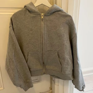 Grå hoodie med dragkedja - Säljer en grå hoodie med dragkedja och huva. Den har två fickor framtill och ribbade muddar vid ärmslut och nederkant. 