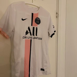 Vit och rosa PSG fotbollströja - Snygg PSG fotbollströja i vitt med rosa och svarta detaljer. Tröjan har korta ärmar och klubbens emblem på bröstet. Perfekt för fans av Paris Saint-Germain!
