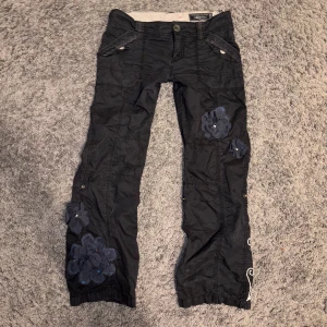 Svarta  byxor med blommiga detaljer - Unika svarta semi-cargobyxor med broderade blommor och applikationer. Byxorna är dekorerade med både stora och små blommor och brodderi. De är lågmidjade och utsvängda. Innerbenslängd 78 cm, tvärs över midjan 42 cm ❤️PRISET ÄR DISKUTERBART!!! ❤️