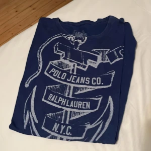 Blå t-shirt från Ralph Lauren - Snygg blå t-shirt från Ralph Lauren med tryck av ett ankare och texten 'Polo Jeans Co. Ralph Lauren N.Y.C.'. Perfekt för en avslappnad stil.