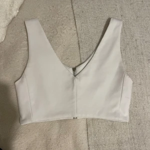 Vit korsettliknande topp med dragkedja - Säljer en stilren vit korsettliknande topp med dragkedja bak .Toppen har en v-ringad design och är ärmlös, vilket gör den perfekt för både vardag och fest. Den är i nyskick och passar bra till olika outfits.