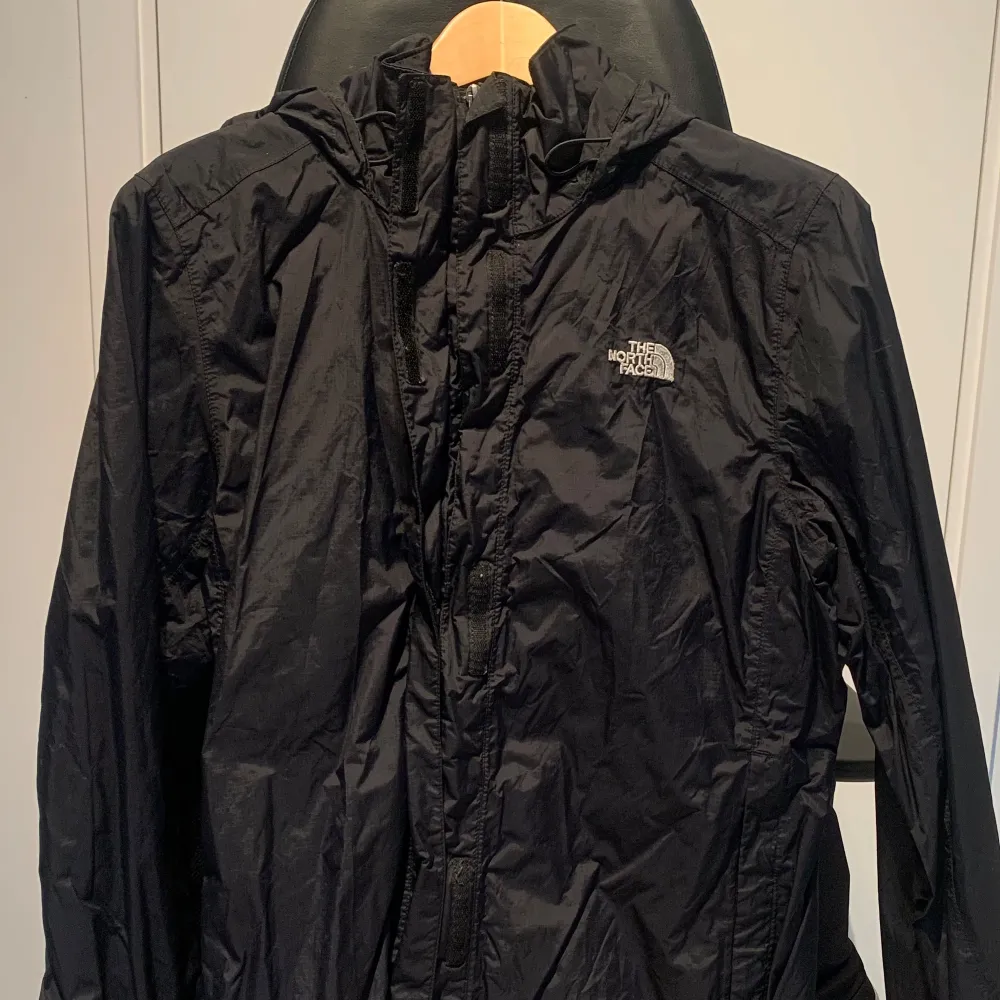 Säljer en svart vindjacka från The North Face. Den är perfekt för blåsiga dagar och har en praktisk huva. Jackan har en dragkedja framtill och är i ett lätt och smidigt material. Den är i bra skick och redo för nya äventyr!. Jackor.