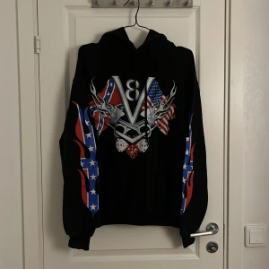 Svart hoodie med V8-motiv - Säljer en svart hoodie med ett coolt V8-motiv på både fram- och baksidan. Den har långa ärmar med ett färgglatt tryck av flammor på sydstatsflaggan. Perfekt för dig som gillar bilar och vill ha en unik stil. Hör av dig om du är intres