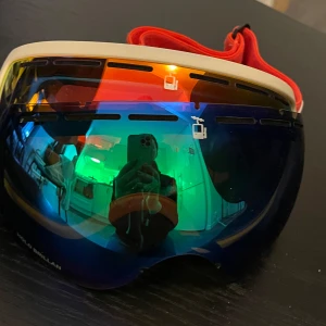 Skidglasögon från House of Hygge - Säljer ett par coola skidglasögon från House of Hygge. De har en spegelblå lins med regnbågseffekt och en vit ram. Det röda bandet är justerbart och har märkets logga. Perfekta för vinterns äventyr i backen!