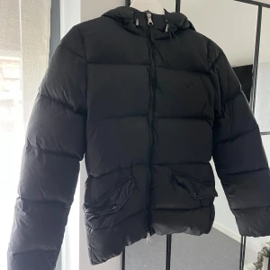 Svart dunjacka från GANT - Säljer en snygg och varm svart dunjacka från GANT. Jackan har en dragkedja framtill och praktiska fickor med knappar. Perfekt för kalla vinterdagar. Den har även en huva! Köpt för något år sen för ca 2000kr 