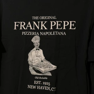 Vintage t-shirt från Port & Company med Frank Pepe-tryck - Säljer en svart t-shirt från Port & Company med ett tryck av Frank Pepe Pizzeria Napoletana. T-shirten har korta ärmar och är tillverkad i bomull. Perfekt för pizzaälskare och fans av klassiska pizzerior!