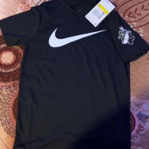 Svart t-shirt från Nike med AIK-logga - Snygg svart t-shirt från Nike med stor vit logga på framsidan och AIK-logga på ärmen. Baksidan har texten 'Allmänna Idrottsklubben' i vitt. Perfekt för fotbollsfans!