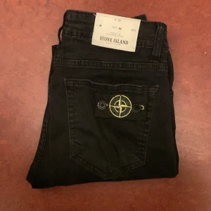 Svarta jeans från Stone Island - Snygga svarta jeans från Stone Island med klassisk femficksdesign och ikonisk logga på bakfickan. Jeansen har en slitning på framsidan för en trendig look. Perfekta för dig som vill ha stil och komfort i ett. Skriv till mig för mer info!😄