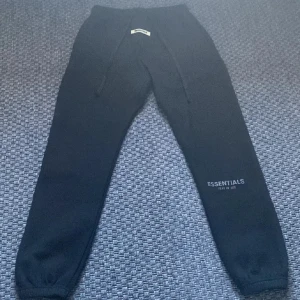 Svarta mjukisbyxor från Essentials - Snygga svarta mjukisbyxor från Essentials Fear of God med elastisk midja och snörning. Perfekta för en avslappnad stil med ribbade muddar vid bensluten och logotyptryck på benet.