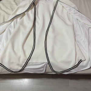 Vit kappa med svarta detaljer - Elegant vit kappa med svarta kantdetaljer. Perfekt för en stilren look. Den har en öppen design utan knappar eller dragkedja.